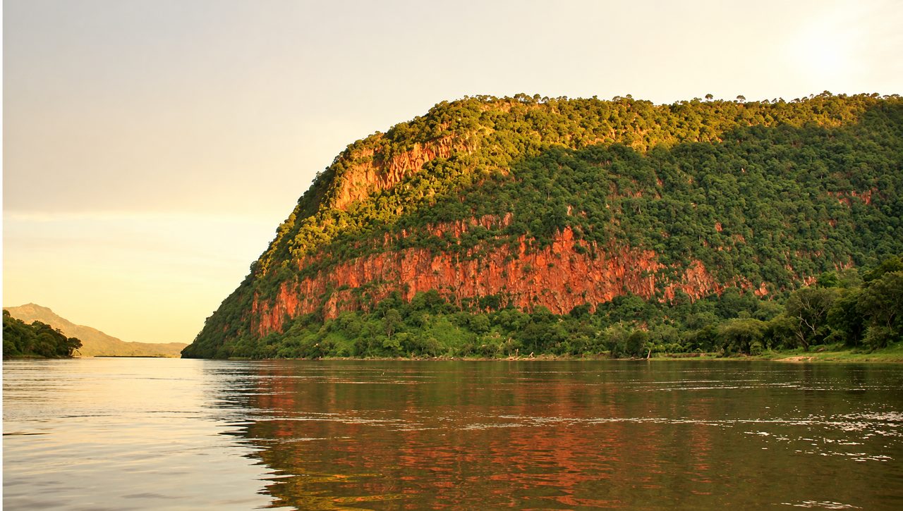 Golden-hour view along the Zambezi, a red-rock ridge rising above calm water
